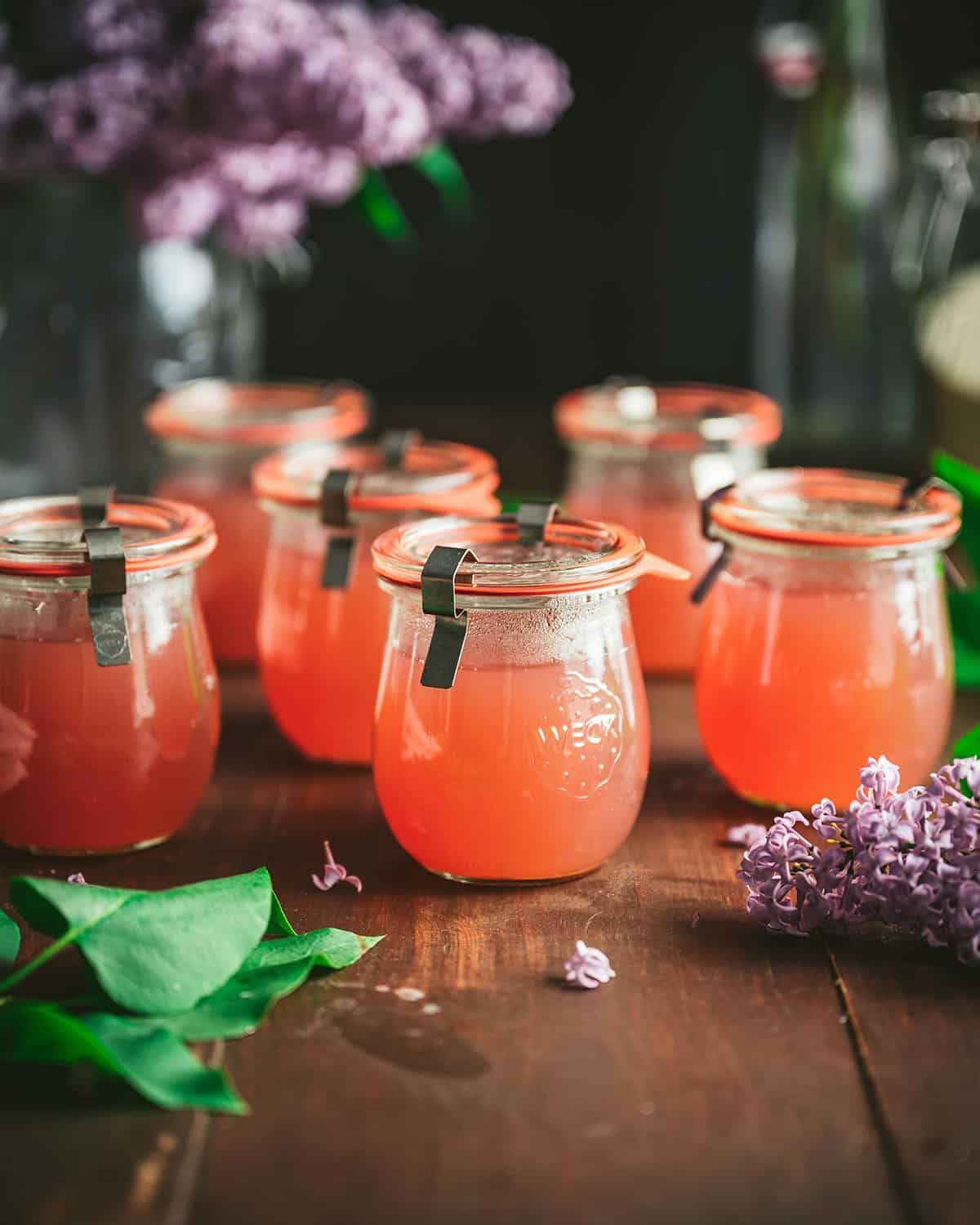 Finished jars of pink lilac jelly on a wood surface surrounded by fresh lilac flowers. 