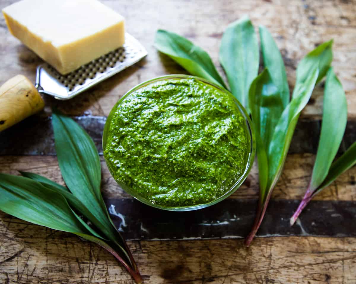 A dish of ramp pesto on a wooden cutting board surrounded by ramp leaves and a block of parmesan and a cheese grater. 