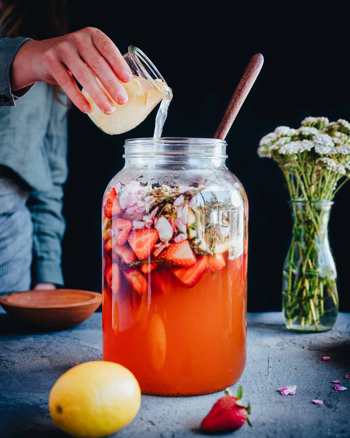 Ginger bug being added to the large jar with soda base, strawberries, rose petals, and yarrow. 