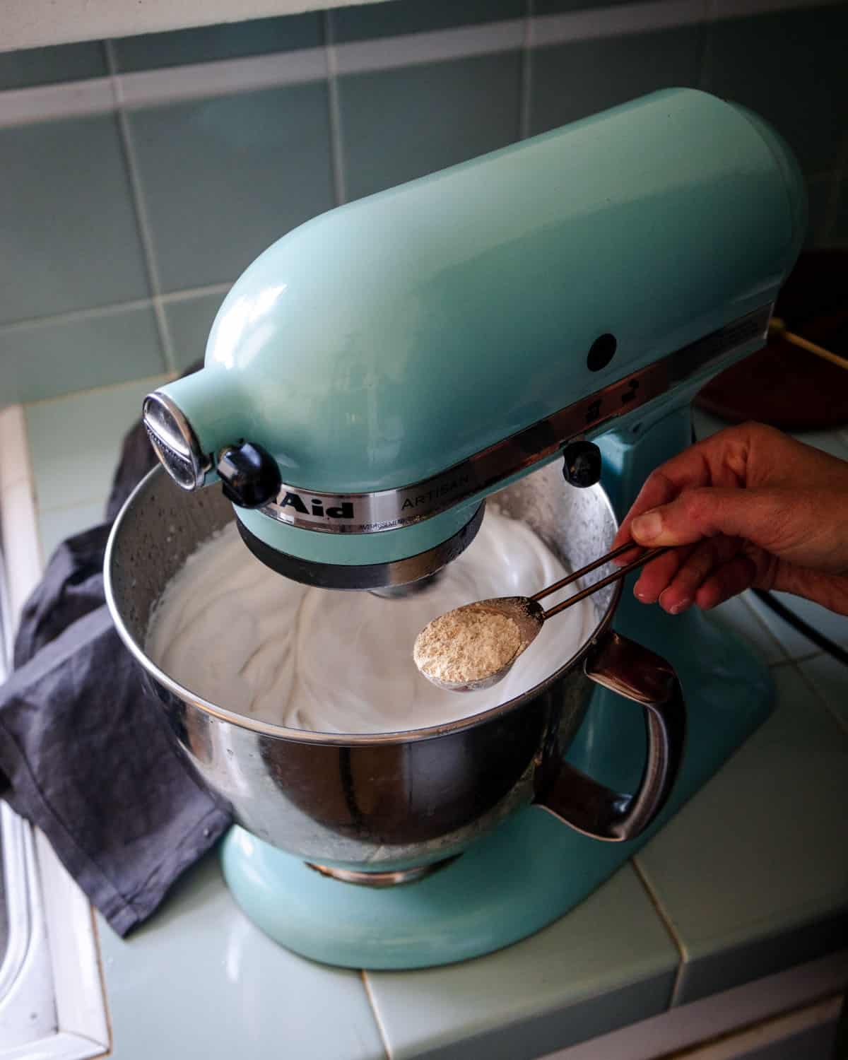 A stand mixer with liquid in it, and marshmallow root powder being added with a spoon. 