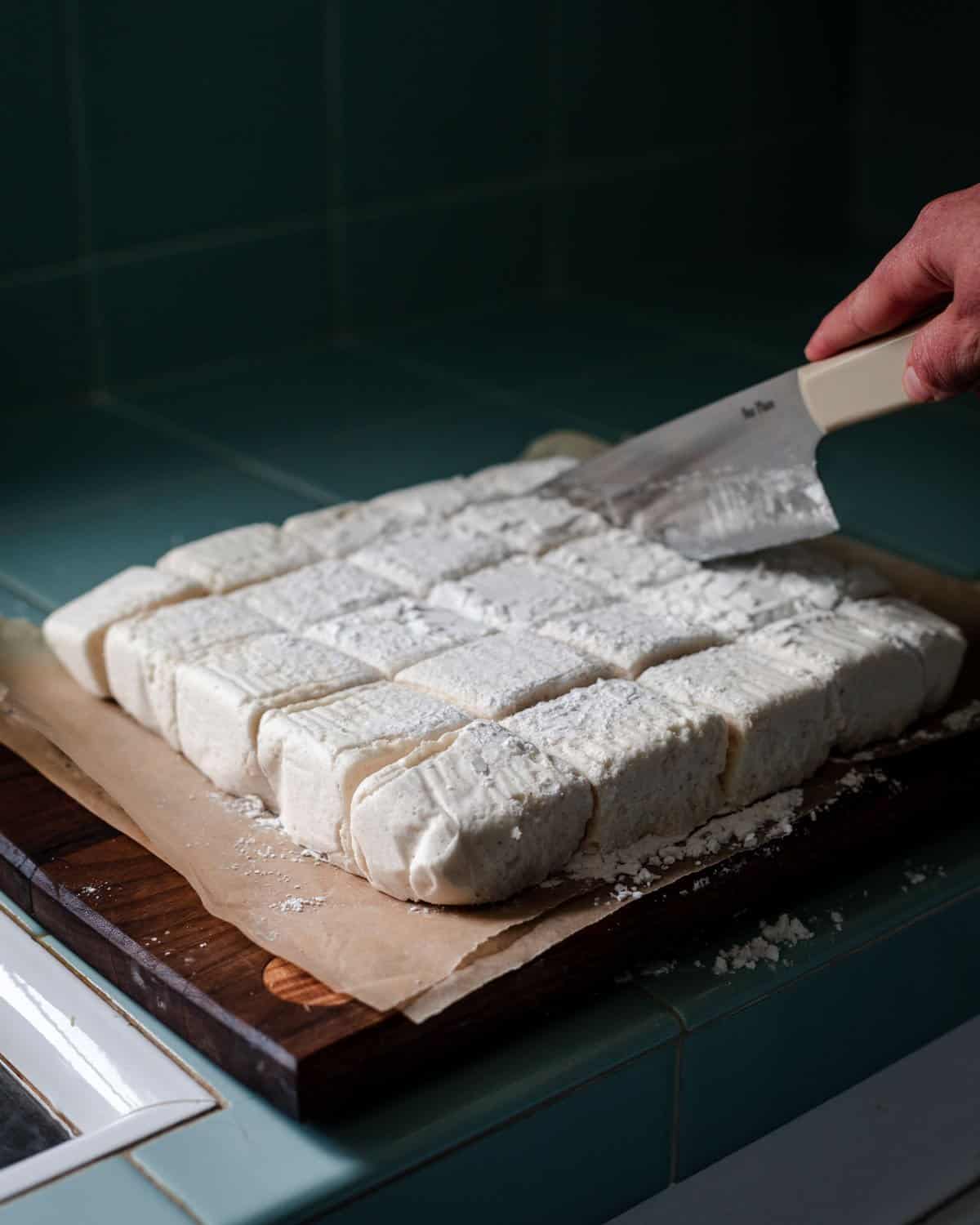 Marshmallows turned onto a cutting board and being cut into squares with a sharp knife. 