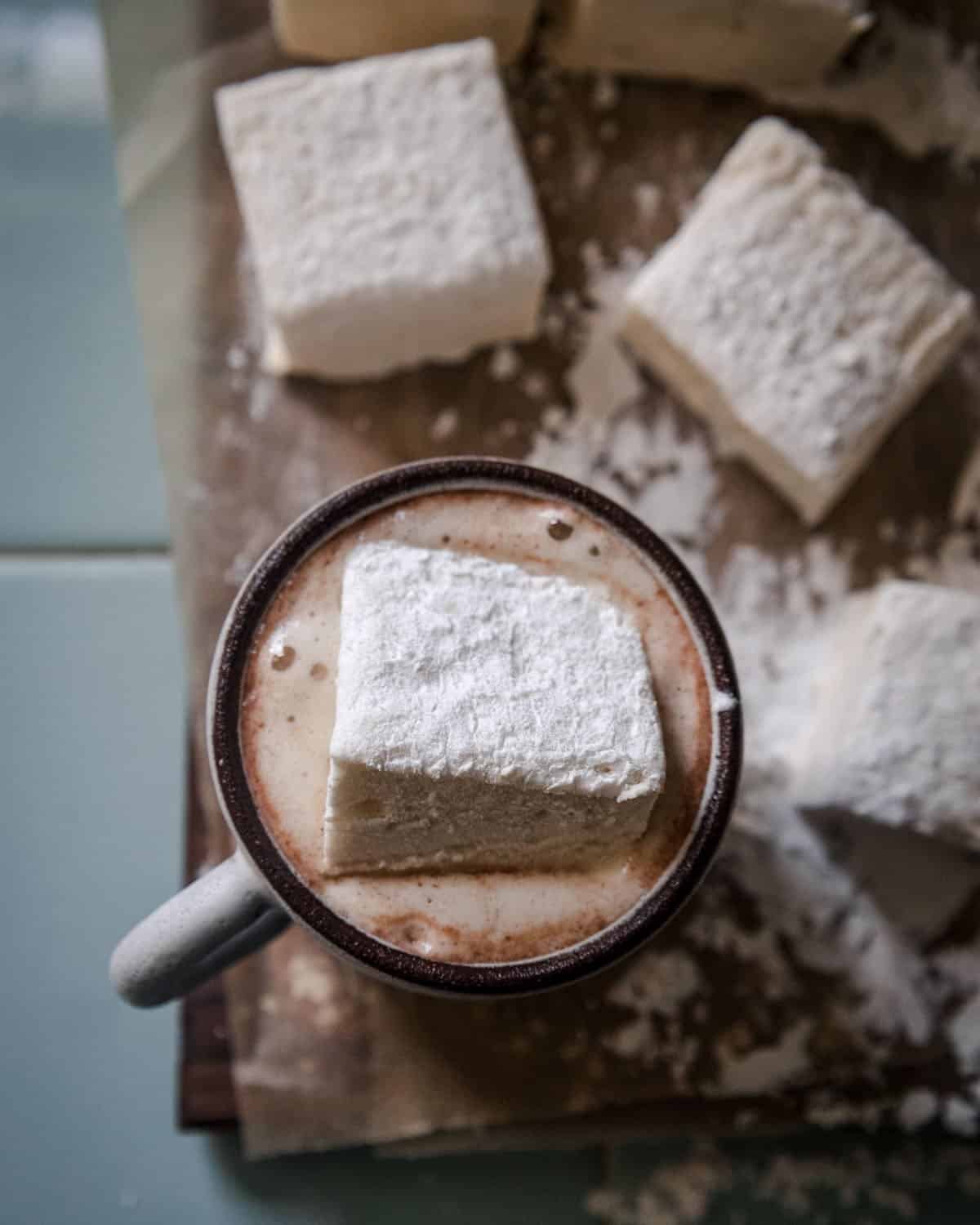 A marshmallow in a mug of hot chocolate, on a cutting board surrounded by other homemade marshmallows. 