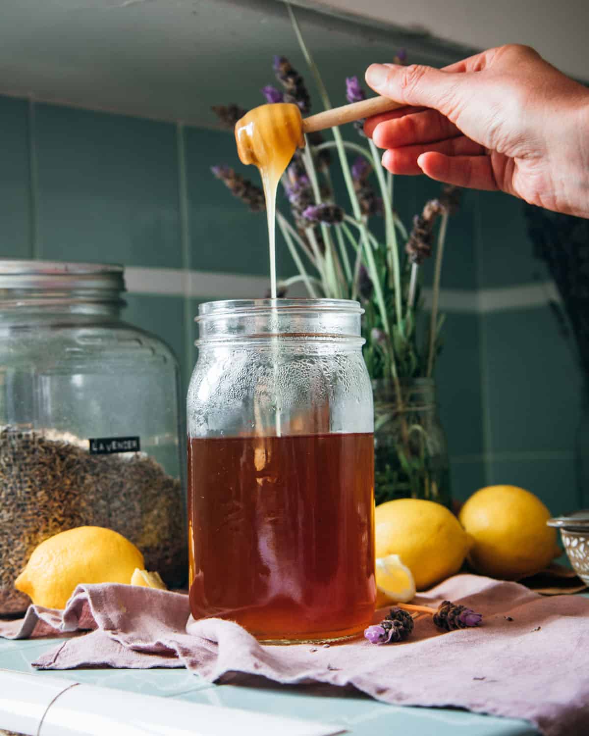 A jar of mostly cooled tea with honey pouring in, surrounded by lemons and a jar of dried lavender flowers. 