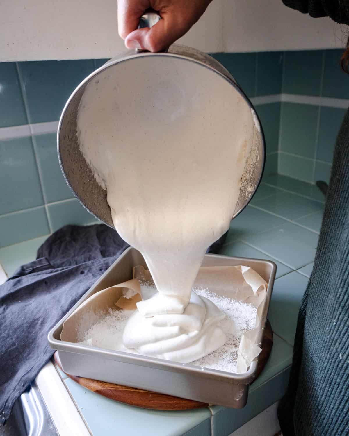 The stand mixer bowl tipped over a pan and pouring the marshmallow fluff into the square pan. 
