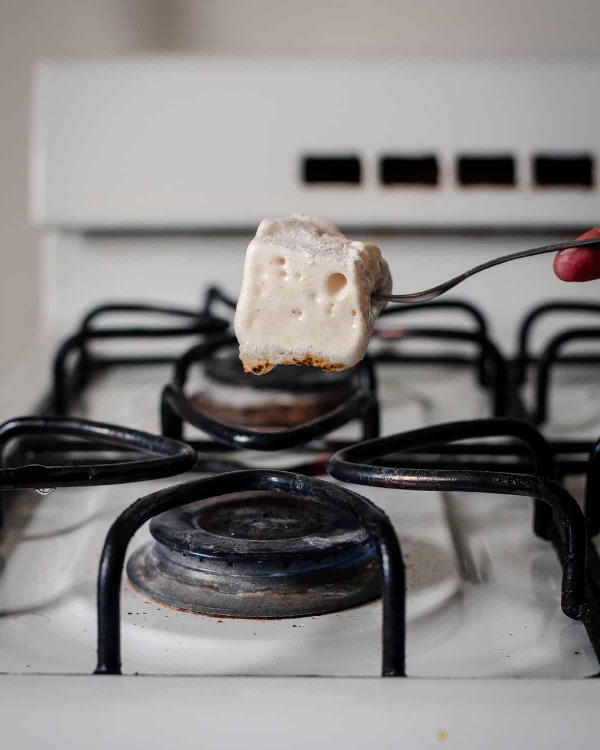 A homemade marshmallow cube on a fork roasting on a gas stovetop. 