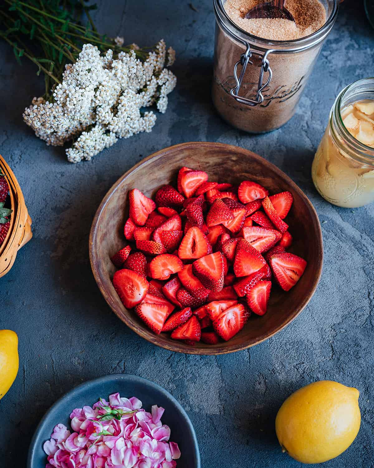 Sliced strawberries in a bowl surrounded by other soda ingredients, a lemon, yarrow, and rose petals.