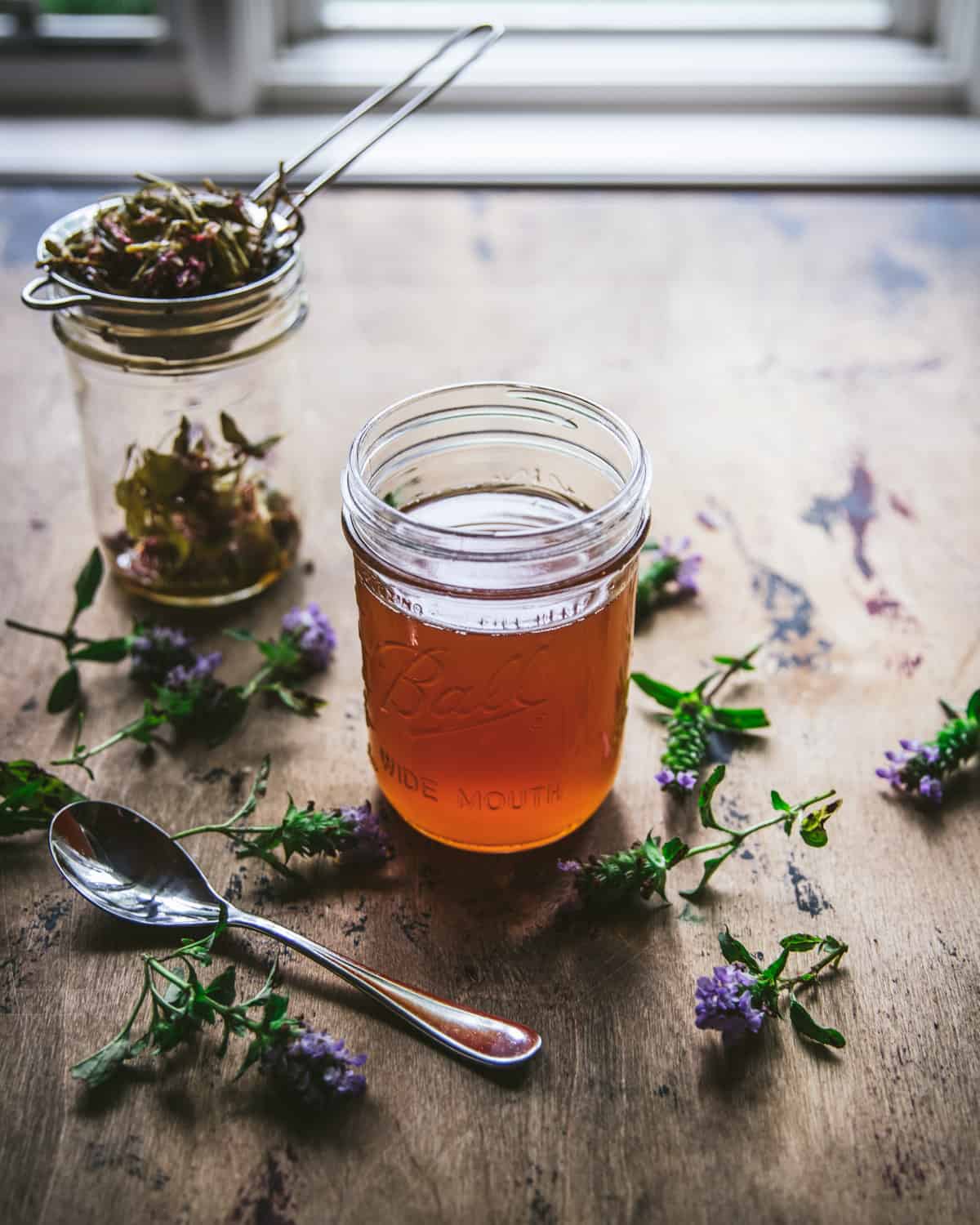 Self-heal oxymel in a jar, surrounded by flowers and a spoon. 