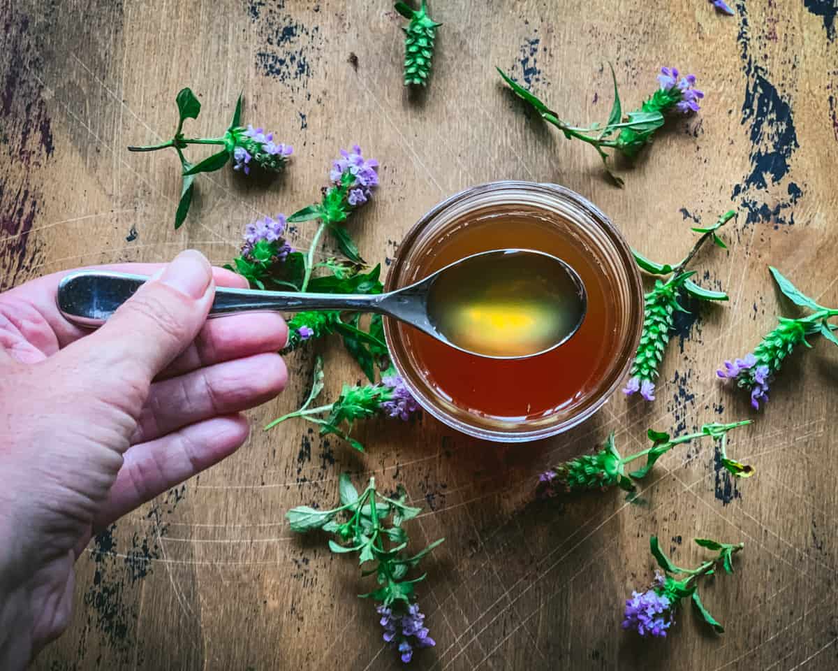 A top view of a jar of self-heal oxymel with a spoon filled lifting out of the jar, and surrounded by self-heal flowers. 