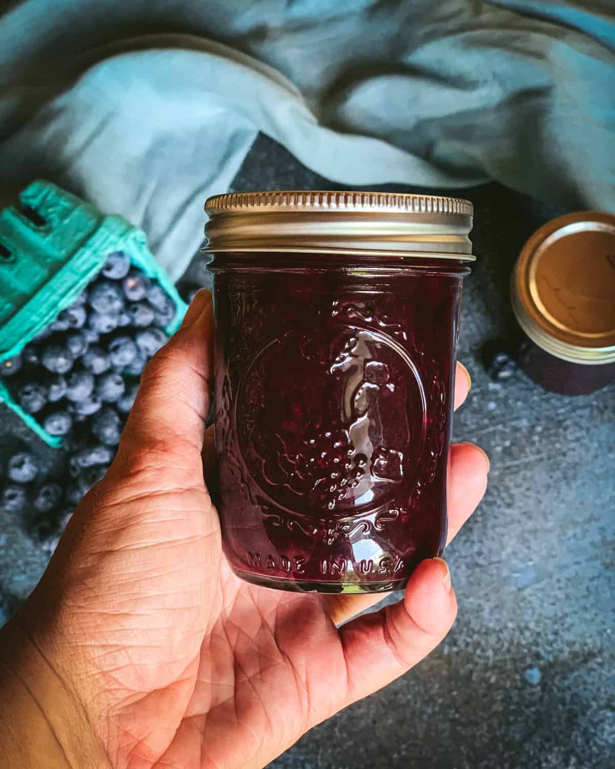 A hand holding blueberry jam in a jar with a lid. 