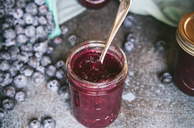 Jar of blueberry jam with a spoon and blueberries surrounding.