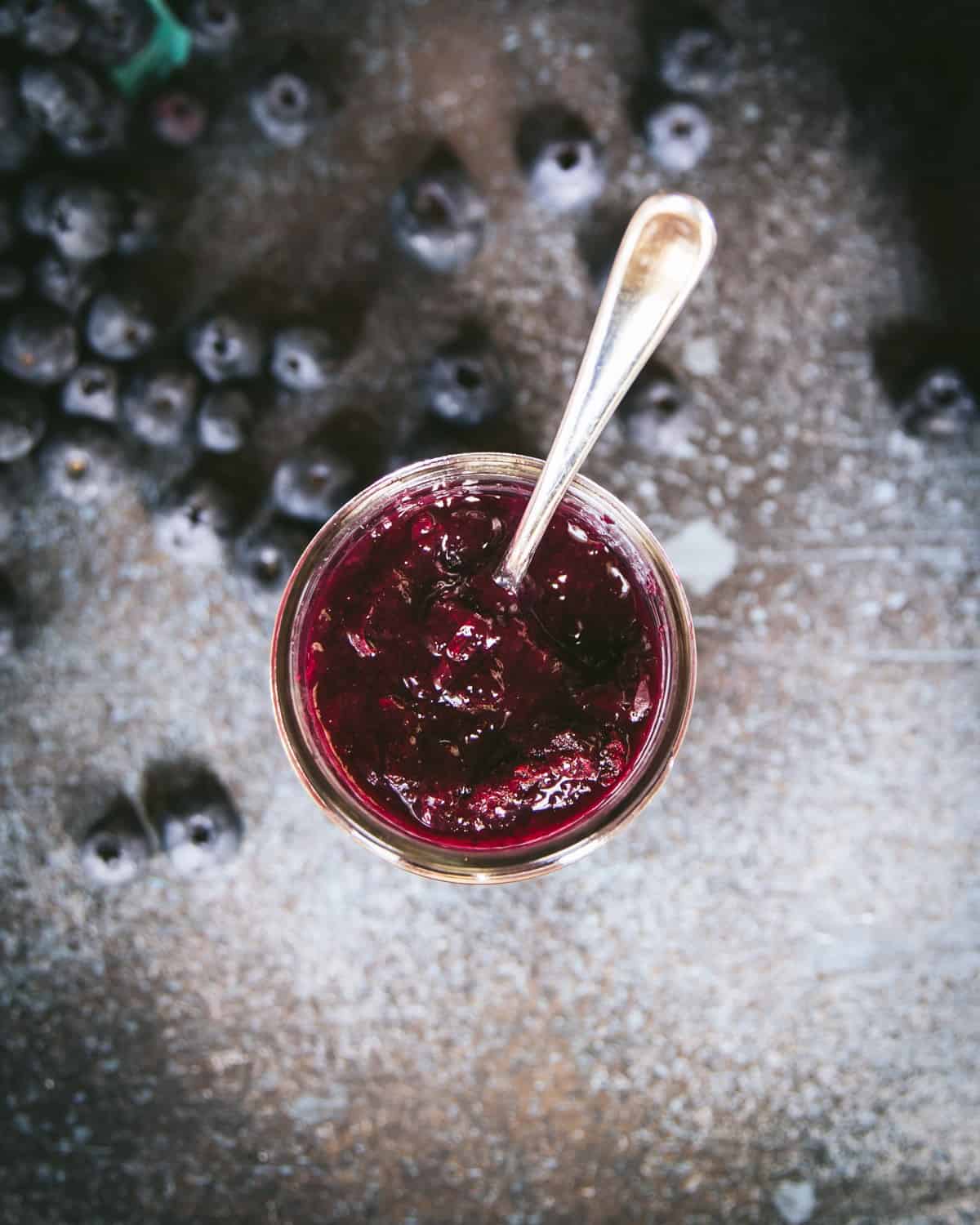 Top view of an open jam jar with a spoon and blueberries surrounding. 