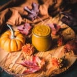 A jar of pumpkin butter surrounded by fall leaves, flowers, and a gourd.