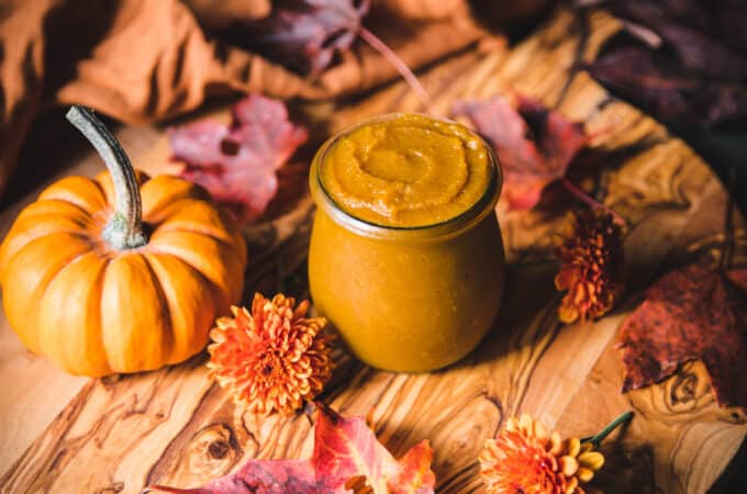 A jar of pumpkin butter surrounded by fall leaves, flowers, and a gourd.