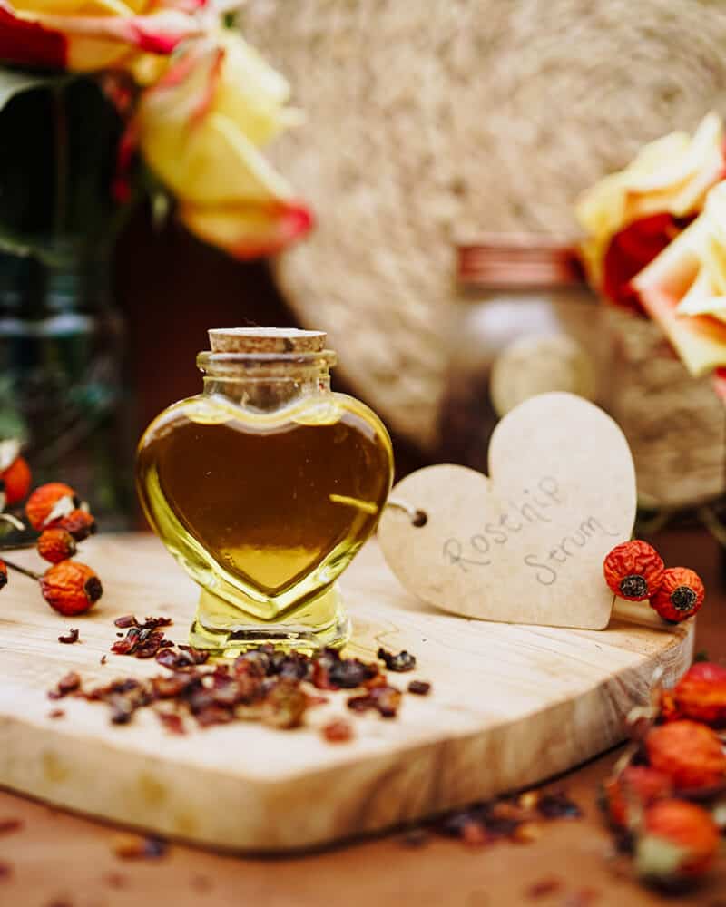 A heart shaped corked bottle with rosehip face oil in it surrounded by fresh and dried rose hips. 