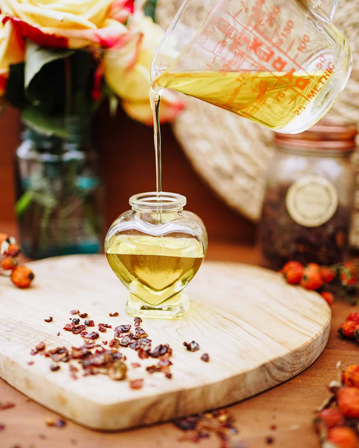 Finished rose hip oil pouring from a measuring cup into a small heart shaped bottle, surrounded by fresh and dried rose hips. 