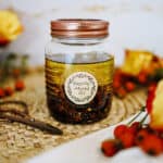 A jar of infusing rose hip oil surrounded by fresh rose hips on a wicker placemat.