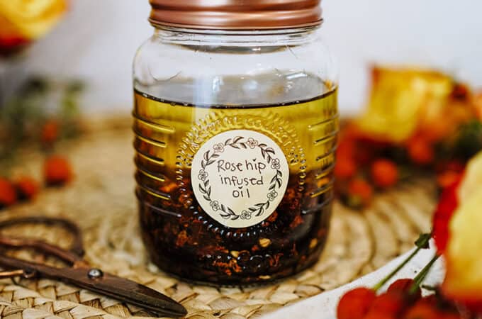 A jar of infusing rose hip oil surrounded by fresh rose hips on a wicker placemat.