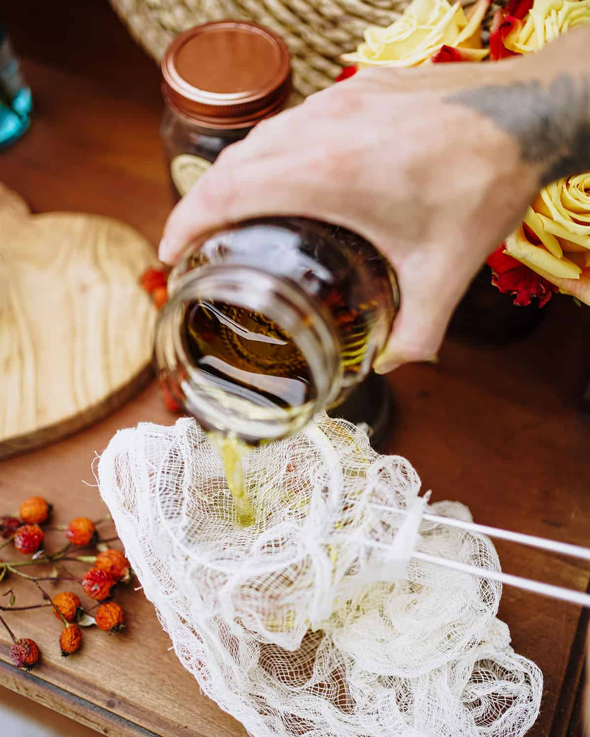 A jar of infused rose hip oil pouring into a mesh strainer with cheesecloth on it to strain out the rose hips, seeds, and hairs. 