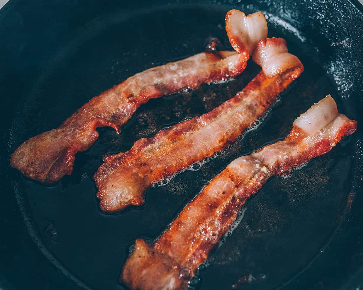 3 strips of bacon frying in a cast iron skillet. 