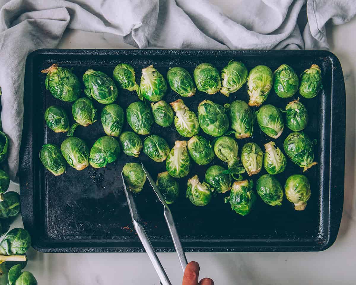 A black sheet pan on a white table, with tongs laying out Brussels sprouts halves cut-side down.