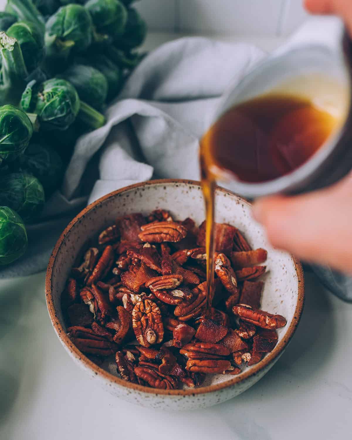 A bowl of chopped bacon and pecan halves with a bowl of maple syrup being drizzled onto it with a stalk of Brussels sprouts in the background.