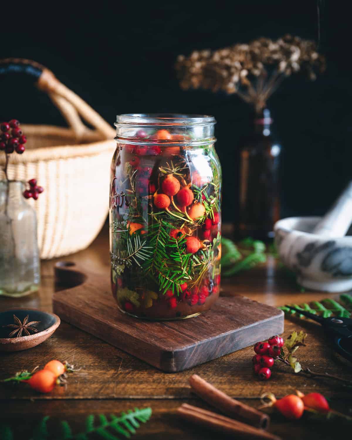 A jar filled with foraged ingredients for mountain vinegar, on a natural wood cutting board surrounded by a basket and other foraged ingredients. 
