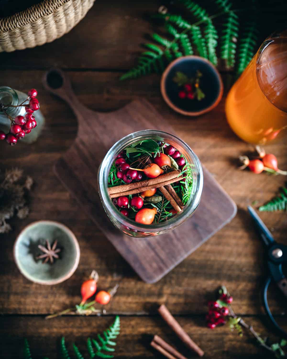 Foraged ingredients in a jar top view with other ingredients surrounding, all on a wooden table and cutting board. 