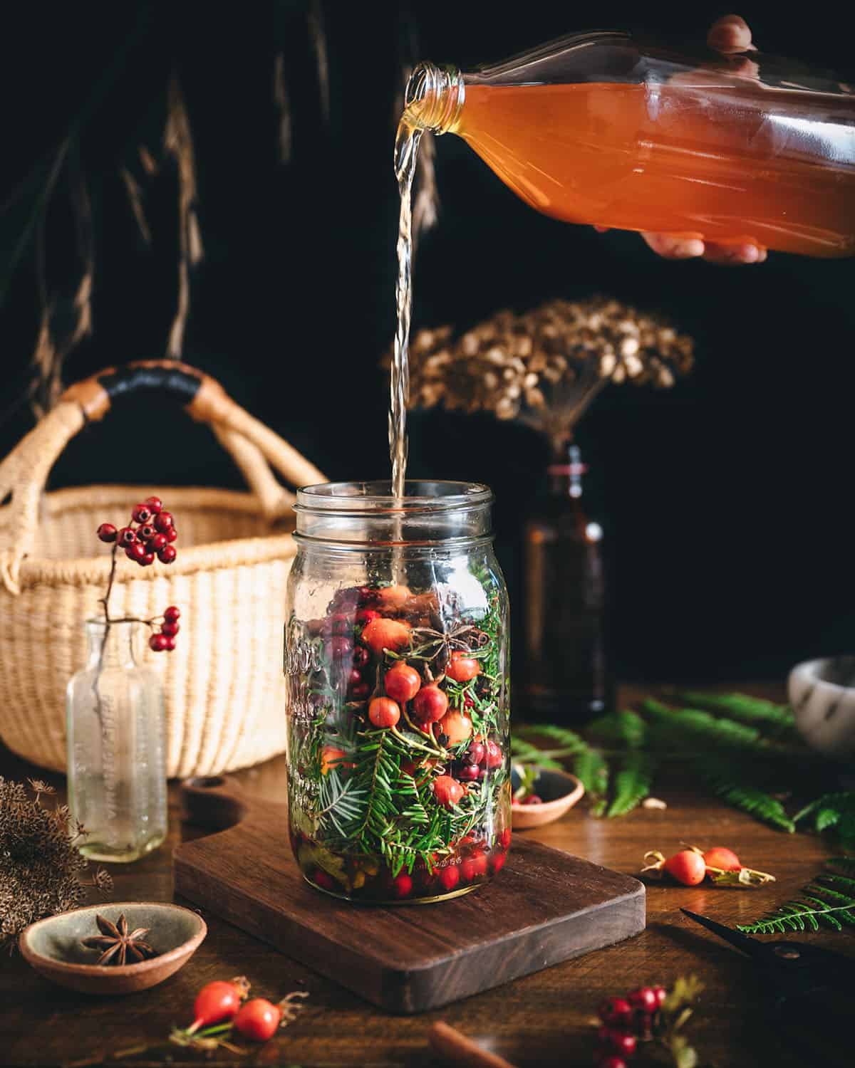 A jar of foraged ingredients in an indoor natural setting, with apple cider vinegar pouring into it to cover the foraged plants and berries. 