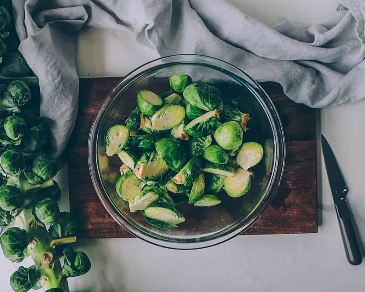 A wooden cutting board on a white table with a knife to the side, and on top sits a bowl of halved Brussels sprouts, with a whole stalk of Brussels sprouts near it.