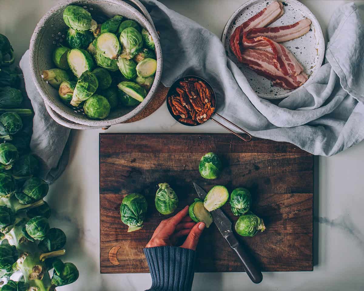 A wooden cutting board with hands cutting Brussels sprouts in half, surrounded by a bowl of bacon, a bowl of chopped Brussels sprouts, and a stalk of Brussels sprouts.