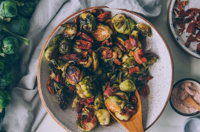 Roasted Brussels sprouts in a bowl with a wooden spoon, surrounded by a white napkin and Brussels sprouts on the stalk.