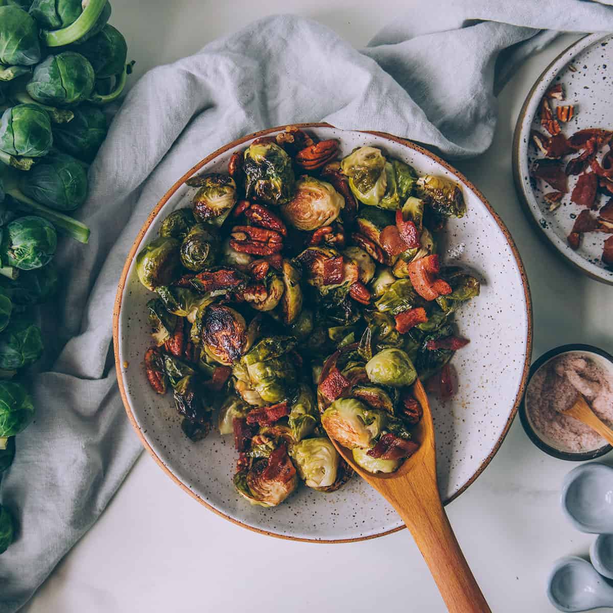 Roasted Brussels sprouts in a bowl with a wooden spoon, surrounded by a white napkin and Brussels sprouts on the stalk. 