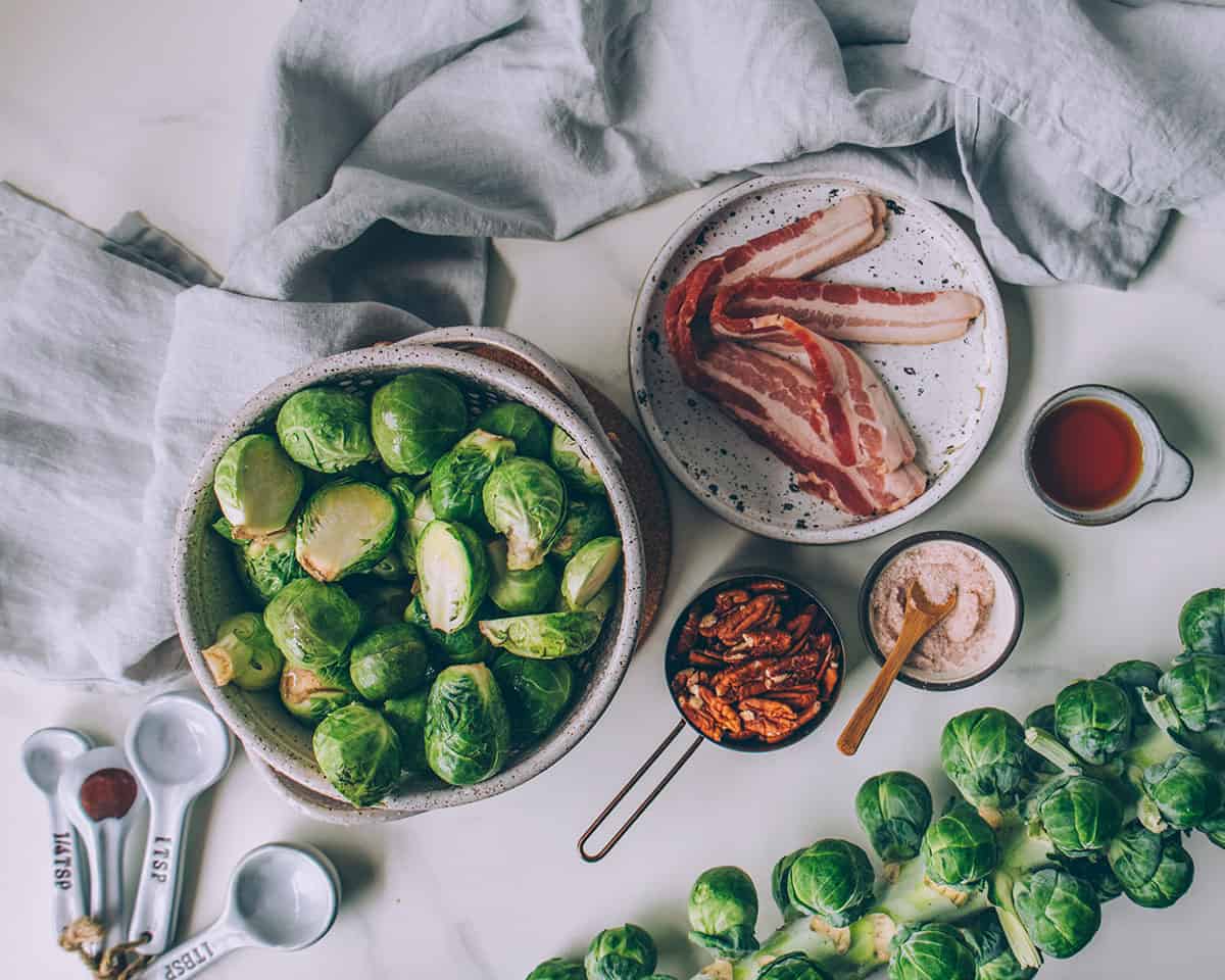 A bowl of halved Brussels sprouts, a stalk of Brussels sprouts, a plate of bacon, and small bowls, measuring cups, and measuring spoons of other ingredients. 