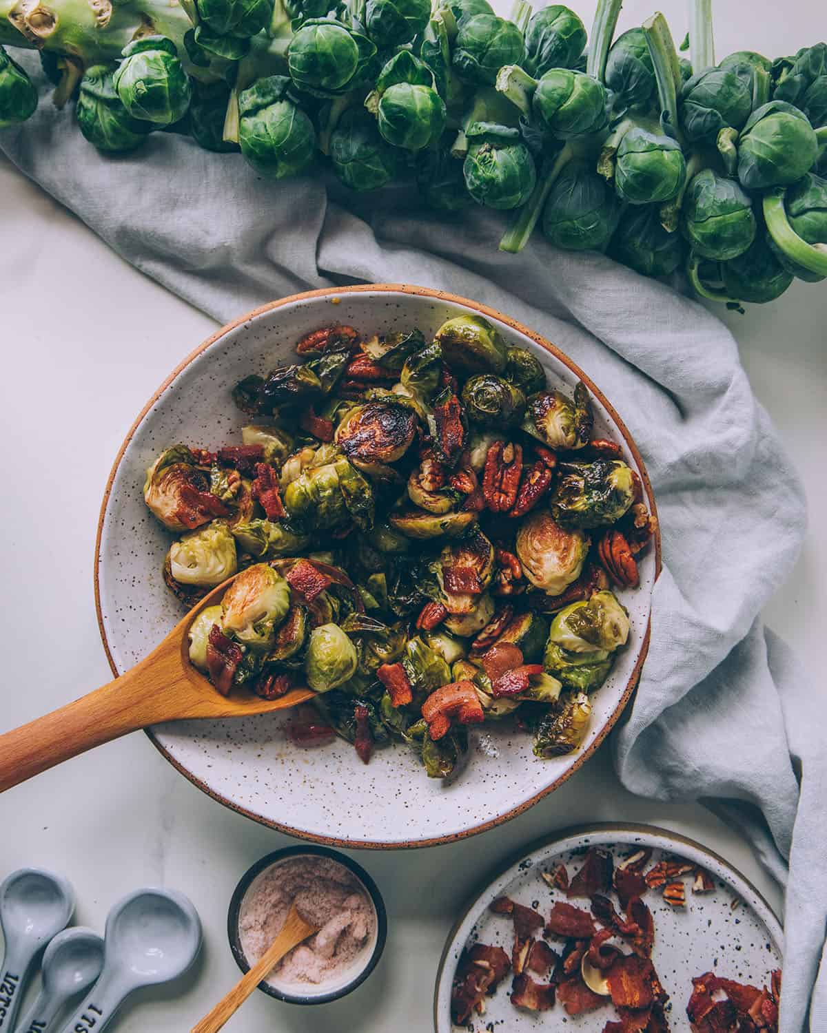 Roasted Brussels sprouts in a bowl with a wooden serving spoon, surrounded by a stalk of Brussels sprouts and other ingredients in bowls. 