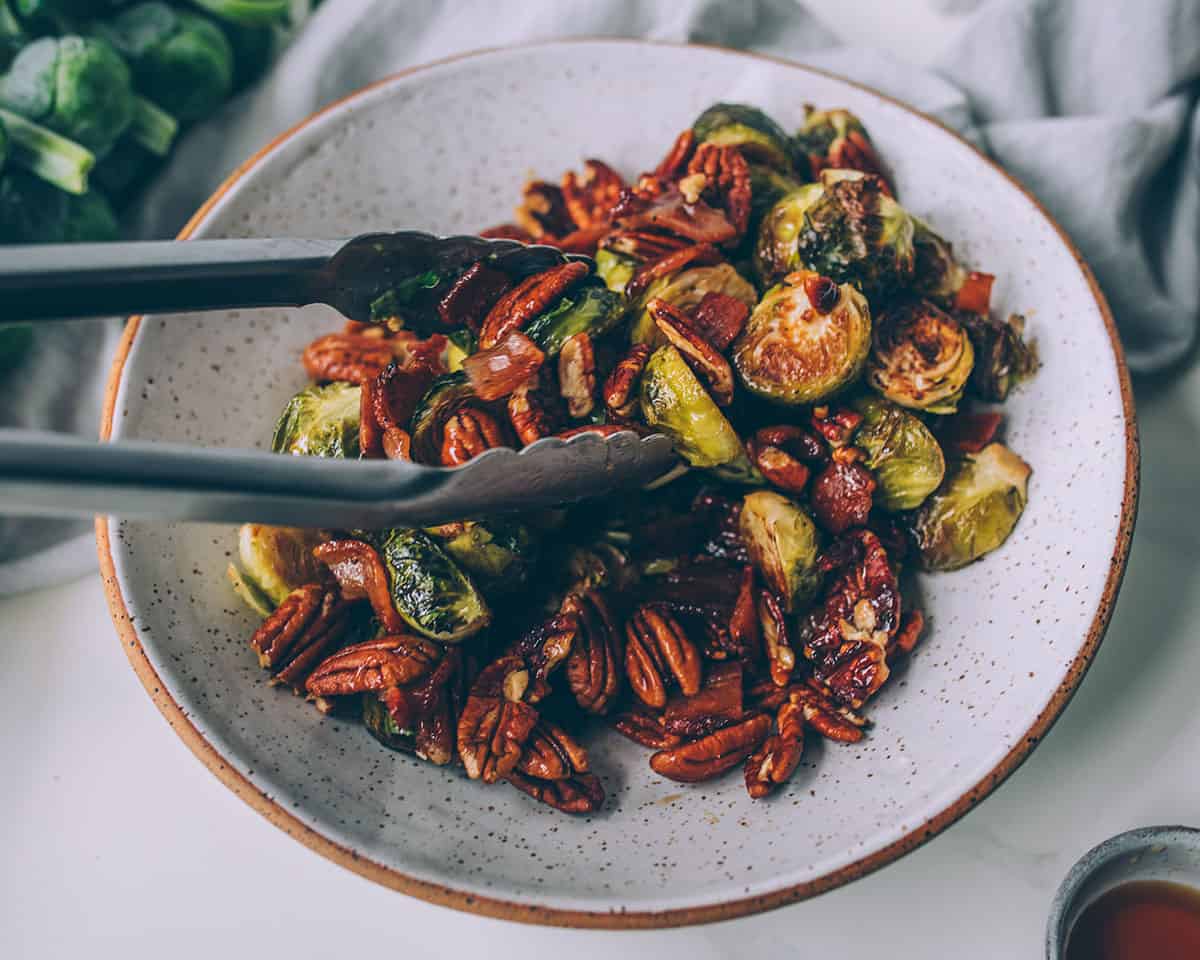 A white bowl with tongs tossing the roasted Brussels sprouts, and pecan, bacon, maple mixture together.
