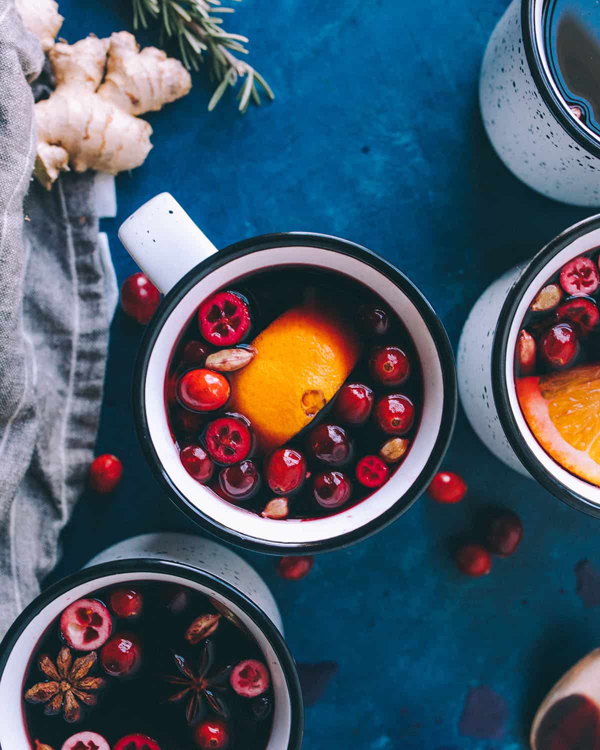 Top view of mugs of mulled wine, garnished with oranges and cranberries, surrounded by fresh ginger and fresh herbs. 