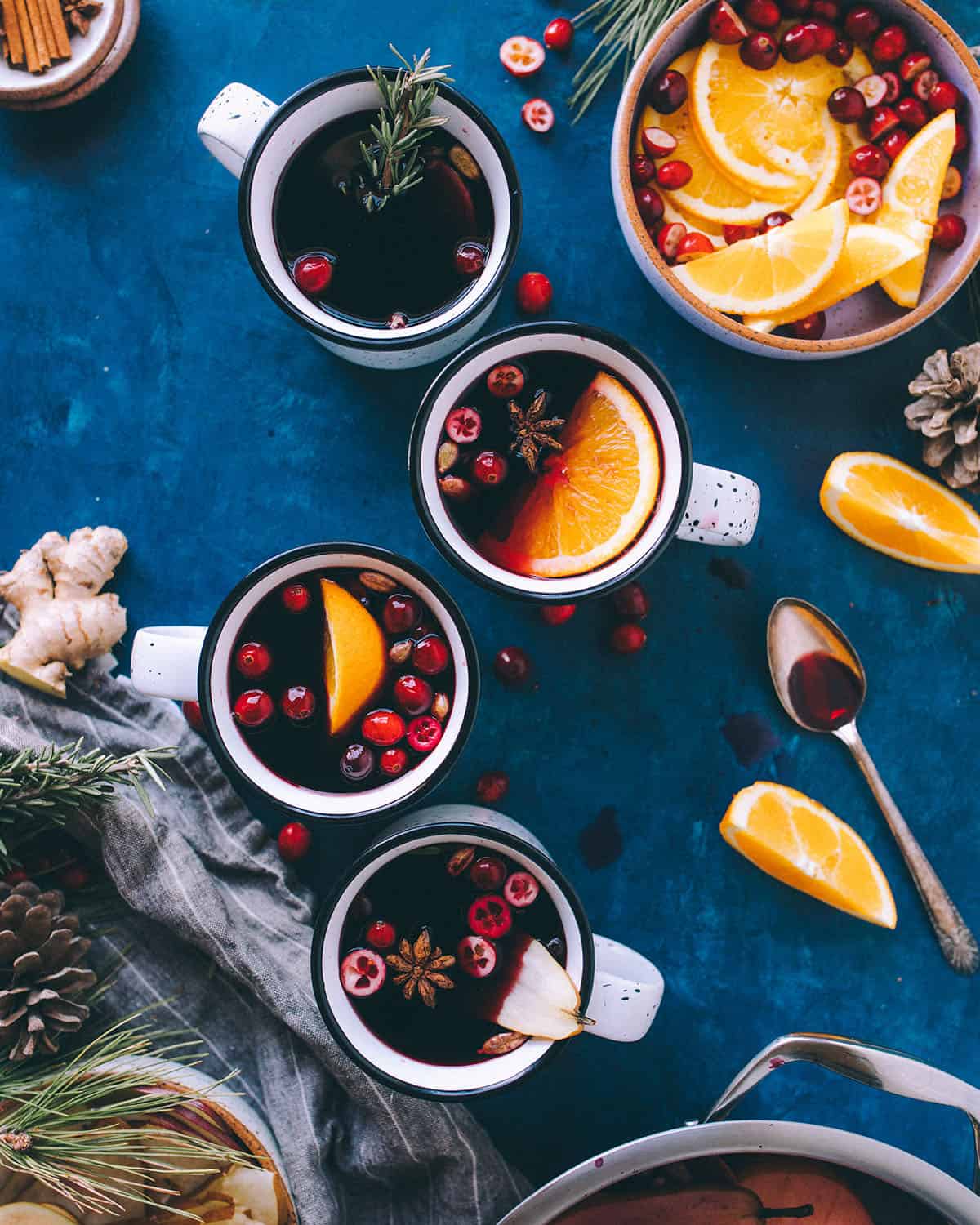 Mulled wine in mugs, garnished with cranberries and orange slices, with a dark background, and a bowl of orange slices to the side. Top view.
