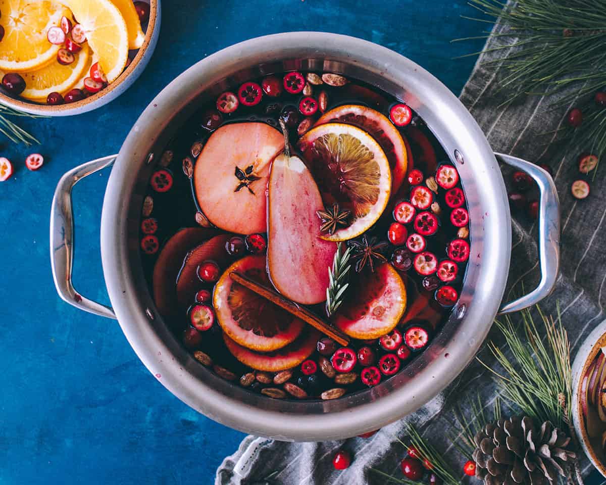 A pot of mulled wine garnished with orange and pear slices, cranberries, and fresh herbs, surrounded by fresh greens and orange slices. Top view.
