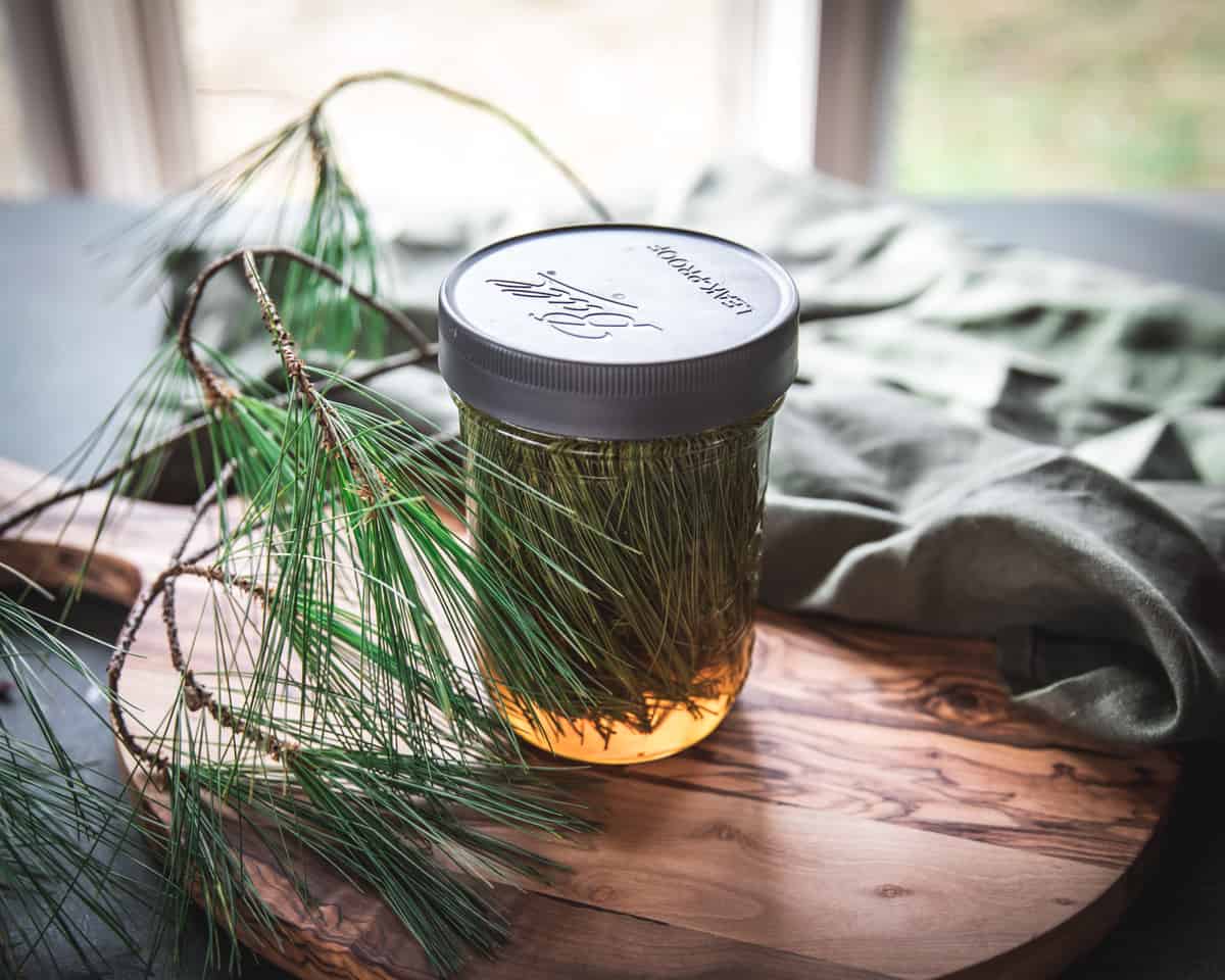 A jar filled with pine needles and honey with a lid, on a wooden cutting board.