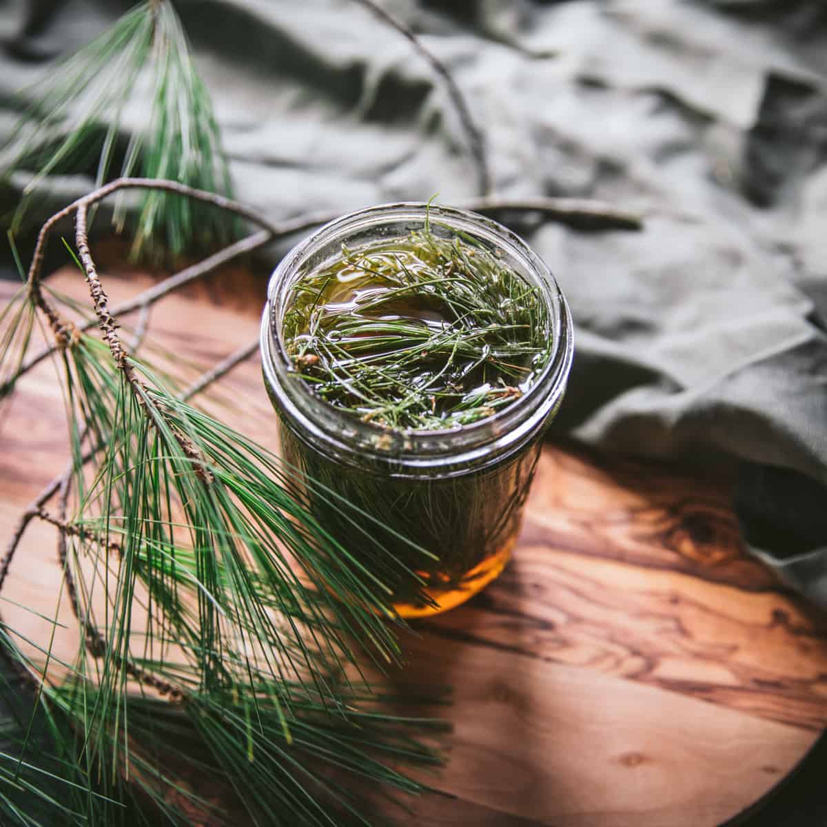 Top view of a jar of honey infusing with pine needles, on a wooden cutting board surrounded by fresh pine fronds.