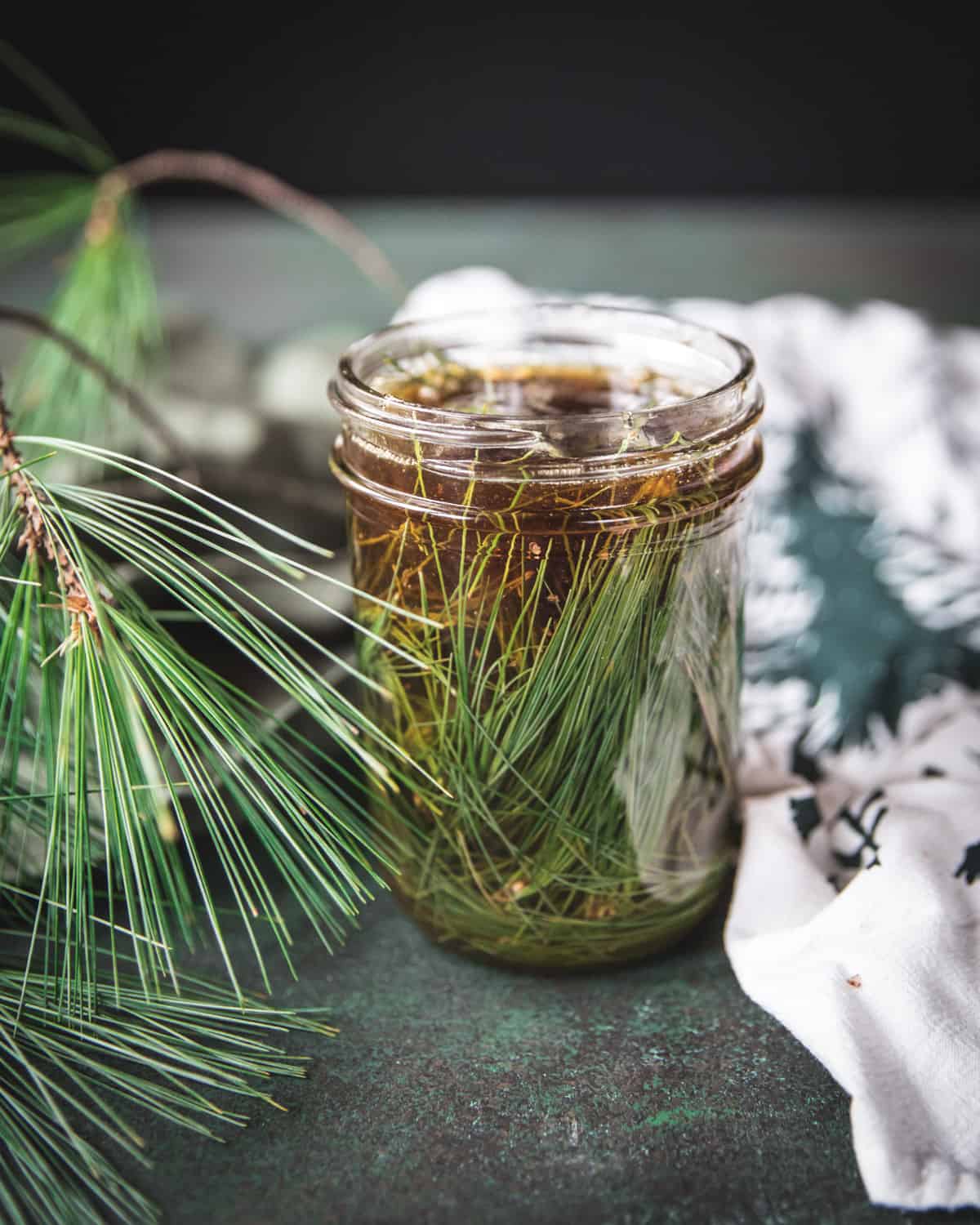 An open jar with pine needles covered in honey, surrounded by fresh pine fronds.
