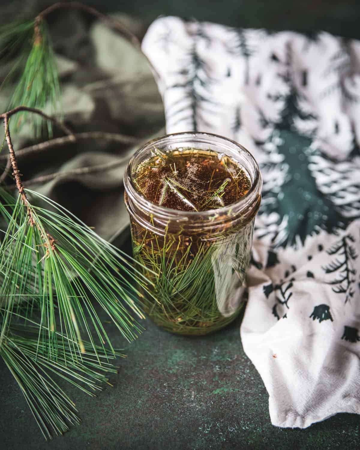 A jar with honey and pine needles, surrounded by fresh pine needles and a tea towel with a green evergreen tree print.
