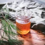 A warm colored pine infused honey in an open jar with a natural cloth green background and pine in the forground,