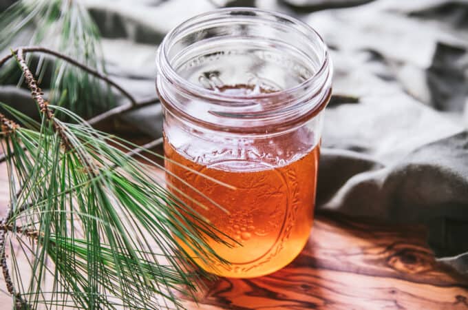 A warm colored pine infused honey in an open jar with a natural cloth green background and pine in the forground,