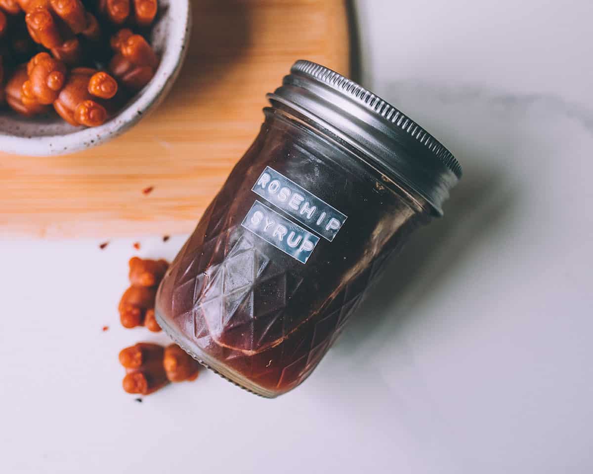 Homemade rosehip syrup in a labeled jar with a lid, surrounded by gummy bears. 