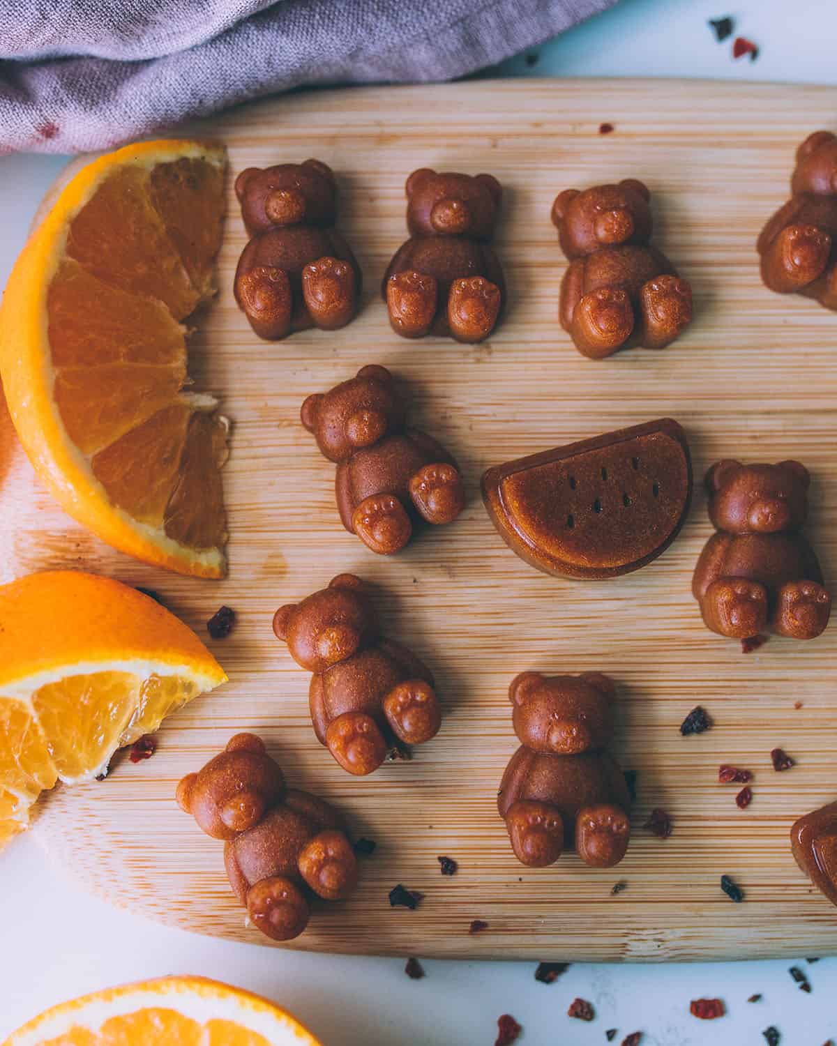 A wooden cutting board with bear shaped vitamin C gummies and orange slices surrounding it. 