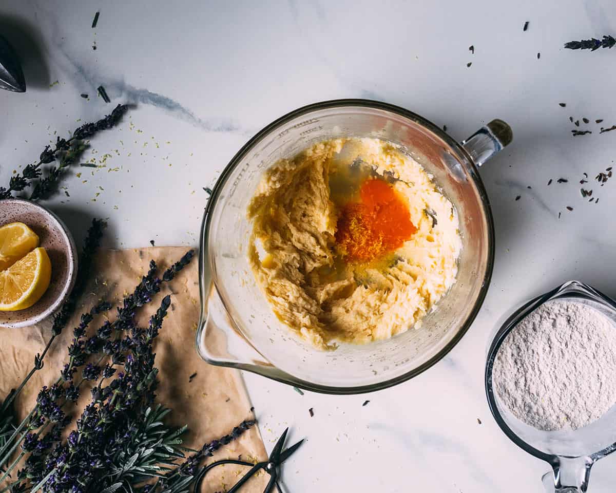 A clear mixing bowl with the dough started and an egg yolk added along with lemon juice, and the arrowroot powder off to the side, surrounded by sliced lemons and fresh lavender.