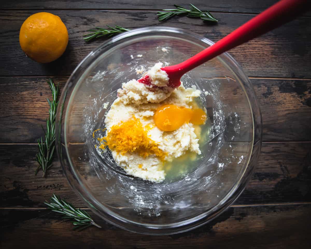 Eggs, lemon juice, and lemon zest added to the clear bowl, stirred with a red rubber spatula. 