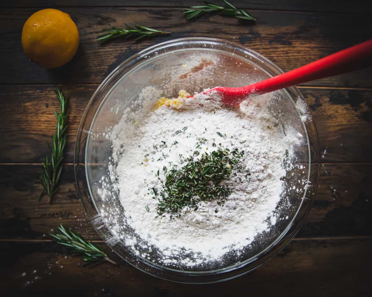 Flour and chopped fresh rosemary added to the bowl, being stirred with a red spatula. 