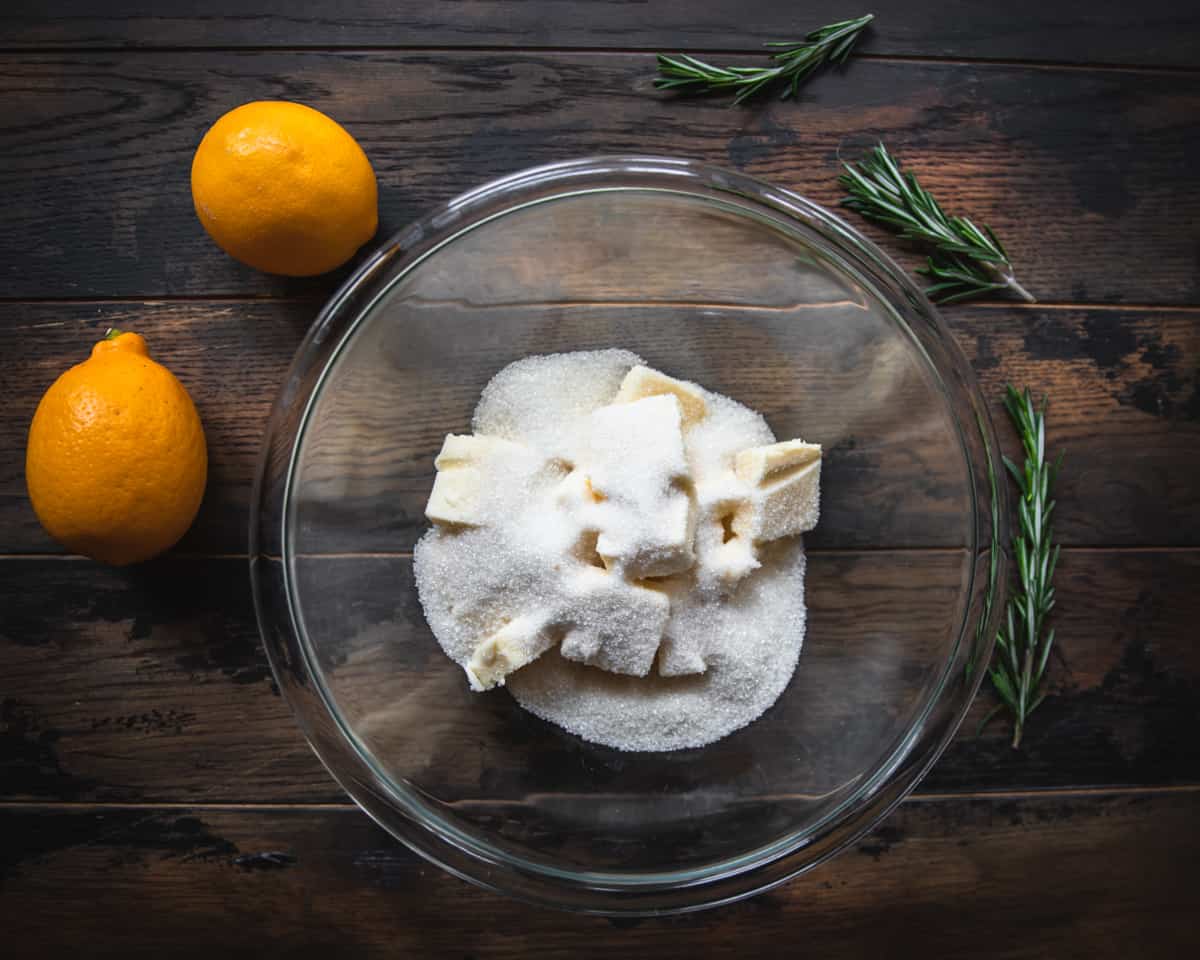 Butter and sugar in a clear bowl on a dark wood surface surrounded by whole lemons and rosemary. 